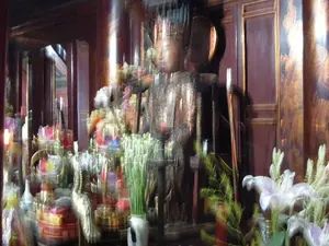 Bức ảnh bị mờ, chụp cận cảnh một ban thờ bên trong một ngôi đền hoặc nơi thờ tự. Ở trung tâm là một bức tượng lớn, có thể bằng đồng, tạc một nhân vật lịch sử hoặc một vị thần, mặc trang phục vua chúa và đội mũ miện. Phía trước tượng là một ban thờ được bày biện trang trọng. Trên ban thờ có rất nhiều đồ cúng lễ, bao gồm hoa tươi như hoa loa kèn trắng, hoa huệ cắm trong lọ, và các lẵng hoa nhiều màu sắc. Ngoài ra còn có nến đang cháy, một con hạc bằng kim loại màu vàng, và các vật phẩm thờ cúng khác được đặt trong các khay, hộp. Phông nền phía sau là các tấm gỗ sơn mài màu nâu sẫm có trang trí hoa văn mạ vàng và các cột gỗ lớn. Không có chữ viết nào có thể đọc được trong ảnh. Khung cảnh mang không khí trang nghiêm, linh thiêng của một nơi thờ cúng.。（日本語訳）。ぼやけた写真で、寺院または礼拝所の内部にある祭壇をクローズアップで撮影したものです。中央には、おそらく青銅製で、王族の衣装を身に着け冠をかぶった歴史上の人物または神を彫った大きな像があります。像の前には、厳かに飾られた祭壇があります。祭壇の上には、白いユリ、花瓶に生けられたチューベローズなどの生花や、色とりどりのフラワーバスケットなど、たくさんのお供え物があります。その他にも、燃えているろうそく、金色の金属製の鶴、そして盆や箱の中に置かれたその他の祭具があります。背景には、金メッキの模様で装飾された濃い茶色の漆塗りの木の板と、大きな木の柱があります。写真の中には、読み取れる文字はありません。その光景は、礼拝所の厳かで神聖な雰囲気を醸し出しています。