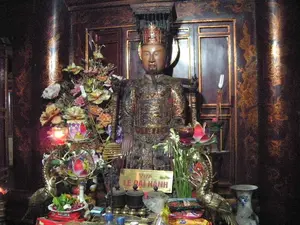 Bức ảnh chụp một ban thờ bên trong một ngôi đền hoặc khu di tích lịch sử. Ở trung tâm là một bức tượng thờ, tạc hình một vị vua đang ngồi, mặc trang phục hoàng gia (long bào) và đội mũ miện. Phía trước tượng có một tấm biển ghi dòng chữ "VUA LÊ ĐẠI HÀNH". Trên ban thờ bày biện nhiều đồ lễ vật. Có thể thấy hoa tươi, hai cây nến lớn hình hoa sen đang cháy sáng, một đĩa đựng trầu cau, các hộp bánh kẹo (có một hộp ghi chữ "BISCUITS"), chai nước và các vật phẩm khác. Hai bên ban thờ có một đôi hạc bằng đồng, mỗi con đỡ một đài nến hình hoa sen. Ngoài ra còn có một lọ hoa bằng sứ và các lư hương nhỏ. Không gian xung quanh được làm bằng gỗ tối màu, với các cột và vách được trang trí hoa văn màu vàng, tạo nên một không khí trang nghiêm, cổ kính.。（日本語訳）。写真は、寺院または史跡の中にある祭壇を写したものです。中央には、王の衣装（龍袍）を身に着け、王冠をかぶって座っている王の姿を彫った祭祀像があります。像の前には「VUA LÊ ĐẠI HÀNH」という文字が書かれた札があります。祭壇の上には多くのお供え物が並べられています。生花、燃えている2本の大きな蓮の形をした蝋燭、キンマとビンロウジを盛った皿、お菓子の箱（「BISCUITS」と書かれた箱が1つ）、水のボトル、その他の品々が見えます。祭壇の両側には一対の銅製の鶴があり、それぞれが蓮の形をした燭台を支えています。その他にも、磁器の花瓶と小さな香炉があります。周囲の空間は暗い色の木材でできており、柱や壁は黄色の模様で装飾され、荘厳で古風な雰囲気を醸し出しています。