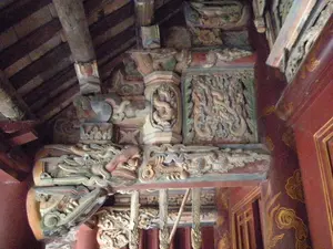 Đây là hình ảnh chụp chi tiết kiến trúc bên trong một công trình cổ, có khả năng là đền, chùa hoặc đình làng. Toàn bộ kết cấu được làm bằng gỗ, với các cột, dầm và mái nhà được chạm khắc vô cùng công phu và tinh xảo. Các tác phẩm điêu khắc chủ yếu mô tả các linh vật trong văn hóa Á Đông, đặc biệt là hình tượng rồng. Có thể thấy rõ một đầu rồng lớn được chạm trổ ở đầu một cây xà ngang, và các bức phù điêu khác khắc họa hình rồng đang uốn lượn. Các cột trụ được sơn màu đỏ, trang trí bằng các họa tiết đám mây màu vàng. Mái nhà có kết cấu bằng gỗ với các thanh xà và kèo lộ ra. Màu sắc của gỗ và các chi tiết trang trí đã cũ và phai mờ, thể hiện sự cổ kính của công trình. Không có hoạt động nào của con người diễn ra và không có chữ viết nào có thể nhận dạng được trong ảnh.。（日本語訳）。これは、古い建造物の内部建築を詳細に撮影した写真で、おそらく寺院、仏塔、または村の集会所（亭）でしょう。構造全体が木でできており、柱、梁、屋根には非常に精巧で緻密な彫刻が施されています。彫刻作品は主に東アジア文化における神聖な生き物を描いており、特に龍の姿が目立ちます。横梁の端に彫られた大きな龍の頭や、うねる龍の姿を描いた他のレリーフがはっきりと見えます。柱は赤く塗られ、黄色い雲の模様で装飾されています。屋根は木造で、梁や垂木が露出しています。木材や装飾の細部の色は古びて色褪せており、建造物の古さを示しています。写真には人の活動は見られず、識別できる文字もありません。