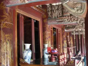 Bối cảnh là không gian bên trong một công trình kiến trúc cổ truyền của Việt Nam, có thể là đền, chùa hoặc một khu di tích lịch sử. Công trình có kết cấu chủ yếu bằng gỗ, nổi bật với các cột nhà to, tròn được sơn son màu đỏ và trang trí bằng các họa tiết rồng, mây uốn lượn được thếp vàng. Hệ thống mái và các vì kèo được chạm khắc gỗ vô cùng tinh xảo và phức tạp, thể hiện kỹ thuật điêu khắc truyền thống. Ở tiền cảnh là một bàn thờ hoặc bàn trưng bày bằng gỗ sẫm màu. Trên bàn có đặt nhiều vật phẩm, bao gồm hai chiếc bình gốm sứ men lam (lộc bình) với hoa văn màu xanh, một bộ chân nến bằng gỗ, một khay gỗ màu đỏ đựng đồ lễ và các hộp gỗ nhỏ hình trụ. Ở phía sau, có một người phụ nữ đang ngồi tại một chiếc bàn khác. Không có chữ viết nào có thể đọc được. Khung cảnh mang đậm nét trang nghiêm, cổ kính và giàu giá trị văn hóa.。（日本語訳）。背景はベトナムの伝統的な建築物の内部空間で、寺院、仏塔、あるいは歴史的遺跡である可能性があります。建物は主に木造で、赤く漆塗りされ、金箔で装飾された龍や渦巻く雲の模様が施された、太く丸い柱が特徴です。屋根の構造と垂木は、伝統的な彫刻技術を示す、非常に精巧で複雑な木彫りが施されています。前景には、濃い色の木製の祭壇または展示台があります。台の上には、青い模様の染付磁器の壺（禄瓶）が二つ、木製の燭台一式、お供え物を乗せた赤い木製の盆、そして小さな円筒形の木箱など、多くの品物が置かれています。後方には、別の机に座っている女性が一人います。読み取れる文字はありません。この光景は、荘厳で古風、そして文化的な価値に富んだ趣を色濃く帯びています。