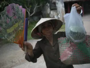 Một người phụ nữ lớn tuổi đội nón lá đang nhìn vào máy ảnh. Bà mặc một chiếc áo sơ mi dài tay màu sẫm. Tay trái bà đang cầm một chiếc quạt giấy xếp có nan màu cam, trên quạt có in hình tranh phong cảnh. Tay phải bà xách một chiếc túi ni lông, bên trong đựng một vật tròn màu xanh có gai, trông giống như một quả na. Bà cũng đang đeo một chiếc túi xách đan bằng nhựa có họa tiết caro màu đỏ và xanh lá cây. Bối cảnh phía sau là không gian ngoài trời với cây cối và một bức tường đá cũ, có thể là một khu di tích hoặc đền chùa.。（日本語訳）。ノンラーをかぶった年配の女性がカメラを見ています。彼女は暗い色の長袖シャツを着ています。彼女の左手はオレンジ色の骨がある折りたたみ式の紙扇子を持っており、扇子には風景画が印刷されています。彼女はまた、赤と緑のチェック柄のプラスチックで編まれたハンドバッグを提げています。後ろの背景は屋外で、木々と古い石垣があり、史跡か寺院かもしれません。