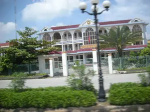 Bức ảnh chụp một tòa nhà lớn, có vẻ là một trụ sở cơ quan hành chính tại tỉnh Ninh Bình. Trên mặt tiền tòa nhà có một tấm biển lớn ghi dòng chữ 'MẶT TRẬN TỔ QUỐC VIỆT NAM HUYỆN...', tuy nhiên tên huyện cụ thể không thể đọc rõ. Tòa nhà có kiến trúc hai tầng, mái ngói màu đỏ, với Quốc kỳ Việt Nam được treo ở ban công tầng hai. Toàn bộ khuôn viên được bao quanh bởi hàng rào sắt màu xanh lá cây có cổng chính. Trong sân có trồng một cây cọ và nhiều cây xanh khác. Phía trước, gần với người chụp ảnh, là một cột đèn đường trang trí và các bụi cây. Ở phía xa bên trái có một cột ăng-ten viễn thông. Bức ảnh có vẻ được chụp từ một phương tiện đang di chuyển do phần tiền cảnh bị nhòe.。（日本語訳）。写真は大きな建物を写しており、ニンビン省にある行政機関の庁舎のようです。建物の正面には「ベトナム祖国戦線...県」と書かれた大きな看板がありますが、具体的な県名ははっきりと読み取れません。建物は2階建ての建築で、赤い瓦屋根を持ち、2階のバルコニーにはベトナム国旗が掲げられています。敷地全体は、正門のある緑色の鉄柵で囲まれています。庭にはヤシの木1本と、その他多くの緑の木々が植えられています。手前、撮影者に近い場所には、装飾的な街灯と植え込みがあります。遠く左側には、通信アンテナ塔があります。前景がぼやけていることから、写真は移動中の乗り物から撮影されたようです。
