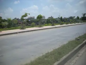 Hình ảnh chụp một con đường nhựa rộng rãi ở một khu vực ngoại ô hoặc nông thôn. Bên cạnh đường là vỉa hè được lát gạch, dọc theo đó là một hàng cây xanh mới trồng còn được chống bằng cọc gỗ. Phía sau vỉa hè là một khu đất thấp hơn có cây cối và thảm thực vật xanh tươi, có thể là một vườn rau. Ở khu vực này, có thể thấy ít nhất hai chiếc máy xúc, một chiếc màu cam và một chiếc màu vàng, cho thấy có một công trình xây dựng hoặc san lấp mặt bằng đang diễn ra. Xa hơn ở phía sau, trên một khu đất cao hơn, là một khu dân cư với nhiều ngôi nhà, một số trong đó dường như đang trong quá trình xây dựng. Bầu trời trong xanh, có vài gợn mây trắng, cho thấy đây là một ngày nắng đẹp. Con đường khá vắng vẻ, không có phương tiện giao thông nào.。（日本語訳）。郊外または田舎の広いアスファルト道路を写した画像。道路の脇にはタイル張りの歩道があり、それに沿って木の杭で支えられた新しく植えられた緑の並木がある。歩道の後ろには、木々や青々とした植物が生い茂る低い土地があり、菜園かもしれない。この地域では、オレンジ色の1台と黄色の1台の少なくとも2台のショベルカーが見られ、建設工事または整地が行われていることを示している。さらに奥の、より高い土地には、多くの家がある住宅地があり、そのうちのいくつかは建設中のようだ。空は青く澄み渡り、白い雲がいくつか浮かんでおり、晴れた美しい日であることを示している。道路はかなり閑散としており、交通車両はない。