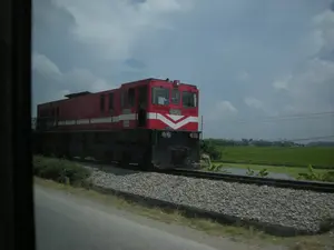 Bức ảnh chụp một đầu máy xe lửa màu đỏ đang ở trên đường ray tại một khu vực nông thôn. Trên thân và mặt trước của đầu máy có ghi rõ số hiệu 'D18E-609'. Đầu máy xe lửa này nằm trên một đường ray rải đá, chạy song song với một con đường nhựa. Bối cảnh phía sau là một cánh đồng lúa xanh bạt ngàn, một số cây chuối và một khu vực mặt nước nhỏ. Trên bầu trời có nhiều mây. Khung cảnh cho thấy một ngày bình thường ở vùng nông thôn, với sự hiện diện của phương tiện giao thông đường sắt và đường bộ xen lẫn với cảnh quan nông nghiệp đặc trưng của Việt Nam.。（日本語訳）。機関車の車体と前面には、「D18E-609」という番号がはっきりと記されています。この機関車は、砂利が敷かれた線路上にあり、アスファルトの道路と平行して走っています。背景には、広大な緑の水田、数本のバナナの木、そして小さな水域があります。空には多くの雲があります。この光景は、ベトナム特有の農業景観に鉄道と道路交通が混在する、田舎のありふれた一日を示しています。