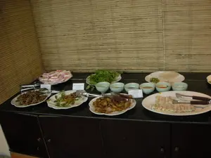 Bối cảnh là một quầy phục vụ thức ăn tự chọn (buffet), có thể ở trong một nhà hàng hoặc khách sạn. Trên một chiếc tủ/quầy màu sẫm, nhiều món ăn đặc sản Việt Nam được bày ra trên các đĩa sứ trắng. Có các tấm biển nhỏ ghi tên món ăn bằng tiếng Việt và tiếng Anh. Các dòng chữ có thể đọc được bao gồm: 'DÊ XÀO LĂN', 'DÊ VIÊN NƯỚNG', và 'CÁ NƯỚNG (Banana grilled / Chich fish)'. Các món ăn được bày biện bao gồm các món từ thịt dê (đặc sản Ninh Bình), cá nướng, một đĩa có các món cuốn, một đĩa thức ăn màu hồng được cắt thành từng miếng, một đĩa rau thơm tươi, bánh tráng để cuốn và nhiều bát nhỏ đựng các loại nước chấm và gia vị. Mỗi đĩa thức ăn đều có kẹp gắp để thực khách tự phục vụ.。（日本語訳）。背景は、レストランかホテルの中にあると思われる、セルフサービスの料理（ビュッフェ）カウンターです。濃い色の戸棚／カウンターの上に、多くのベトナム名物料理が白い磁器の皿に並べられています。料理名がベトナム語と英語で書かれた小さな札があります。読み取れる文字には、「DÊ XÀO LĂN」、「DÊ VIÊN NƯỚNG」、そして「CÁ NƯỚNG (Banana grilled / Chich fish)」が含まれます。並べられている料理には、ヤギ肉料理（ニンビン省の名物）、焼き魚、巻き物料理が乗った一皿、ピンク色で一切れずつにカットされた料理が一皿、新鮮な香草が一皿、巻くためのライスペーパー、そして様々な種類のつけダレや調味料が入った多くの小鉢が含まれます。それぞれの料理皿には、客が自分で取り分けられるようにトングが添えられています。