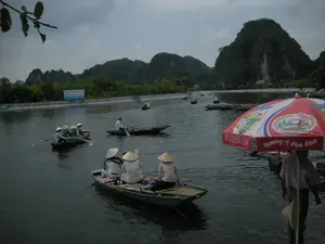 Khung cảnh diễn ra trên một con sông hoặc hồ rộng tại một khu du lịch ở Ninh Bình, được bao quanh bởi những dãy núi đá vôi hùng vĩ và cây cối xanh tươi. Bầu trời u ám và có nhiều mây. Trên mặt nước có rất nhiều thuyền đò nhỏ (ghe) đang chở khách du lịch. Nhiều người trên thuyền, bao gồm cả người chèo thuyền và du khách, đang đội nón lá. Hoạt động chính là du ngoạn bằng thuyền để ngắm cảnh. Một số người dân địa phương đang chèo thuyền đưa du khách đi tham quan. Ở phía trước bên phải, có một chiếc ô lớn màu đỏ và trắng của thương hiệu 'WONDERFARM' với dòng chữ 'Hương vị độc đáo', có thể thuộc về một người bán hàng trên sông. Ở phía xa, bên kia bờ sông có một tấm biển quảng cáo lớn màu xanh lam, trên đó có thể đọc được dòng chữ 'DU LỊCH NINH BÌNH', phần còn lại của văn bản không rõ ràng.。（日本語訳）。ニンビンのある観光地で、広い川または湖の上で繰り広げられる光景。雄大な石灰岩の山々と青々とした木々に囲まれている。空はどんよりとしており、雲が多い。水面には、観光客を乗せた多くの小さな手漕ぎボート（ghe）がある。船頭や観光客を含む、船に乗っている多くの人々がノンラー（菅笠）をかぶっている。主なアクティビティは、景色を眺めるためのボートクルーズである。何人かの地元の人々がボートを漕いで観光客を案内している。右手前には、「WONDERFARM」ブランドの赤と白の大きな傘があり、「Hương vị độc đáo（ユニークな味わい）」という文字が書かれている。これは水上の物売り業者のものかもしれない。遠くの対岸には大きな青い看板があり、「DU LỊCH NINH BÌNH（ニンビン観光）」という文字が読み取れるが、残りのテキストは不明瞭である。