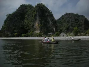 Bối cảnh là một con sông rộng ở khu vực Ninh Bình, được bao quanh bởi những dãy núi đá vôi hùng vĩ, cây cối xanh tươi. Dọc theo bờ sông có một bờ kè bằng đá. Trên mặt nước có hai chiếc thuyền nan (ghe). Chiếc thuyền ở gần hơn đang chở ba du khách và một người phụ nữ địa phương chèo thuyền. Người phụ nữ này đội nón lá và mặc áo màu vàng. Các du khách đang ngồi trên thuyền để tham quan, ngắm cảnh. Ở phía xa hơn, có một chiếc thuyền nan khác với một người cũng đang chèo thuyền và đội nón lá. Trên bờ, có một tấm biển chỉ dẫn màu xanh lá cây với chữ trắng, nhưng chữ viết khó đọc rõ, có vẻ như bắt đầu bằng chữ 'ĐỀN'. Ngoài ra còn có một công trình kiến trúc nhỏ màu trắng và một ngôi nhà mái lá.。（日本語訳）。背景はニンビン地方の広い川で、雄大な石灰岩の山々と青々とした木々に囲まれている。川岸に沿って石造りの堤防がある。水面には2艘の手漕ぎボート（ゲ）がある。より近くにあるボートには、3人の観光客と、船を漕いでいる1人の地元の女性が乗っている。この女性はノンラーをかぶり、黄色い服を着ている。観光客たちはボートに座って観光し、景色を眺めている。さらに遠くには、別の手漕ぎボートが1艘あり、1人の人が乗って船を漕ぎ、ノンラーをかぶっている。その他にも、小さな白い建造物と茅葺き屋根の家が1軒ある。