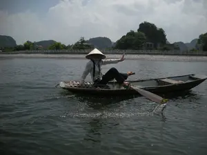 Bối cảnh là một con sông hoặc kênh nước tại một khu vực có nhiều núi đá vôi, có khả năng là một địa điểm du lịch ở Ninh Bình. Một người đang ngồi trên một chiếc thuyền nhỏ (ghe), đội nón lá, mặc áo dài tay và quần sẫm màu. Người này đang chèo thuyền bằng một kỹ thuật đặc biệt là dùng chân để đẩy mái chèo, một kiểu chèo thuyền đặc trưng của vùng này. Một tay của người đó đang giơ lên như đang vẫy chào. Chiếc thuyền có một tấm phên đan bằng tre để ngồi. Một chiếc thuyền trống khác được buộc vào bên cạnh. Ở hậu cảnh, có một bờ kè bằng đá chạy dọc theo bờ sông, phía sau là cây cối và một vài ngôi nhà. Bầu trời có mây và nắng.。（日本語訳）。背景は、多くの石灰岩の山がある地域にある川または水路で、ニンビンの観光地である可能性があります。一人の人が小さなボート（ghe）に座り、ノンラーをかぶり、長袖のシャツと暗い色のズボンを着用しています。この人は、この地域特有の漕ぎ方である、足を使ってオールを押すという特別な技術でボートを漕いでいます。その人の片手は、挨拶をしているかのように上がっています。ボートには座るための竹で編んだ敷物があります。もう一艘の空のボートが横に結び付けられています。後景には、川岸に沿って続く石の護岸があり、その後ろには木々や数軒の家があります。空は雲があり、晴れています。