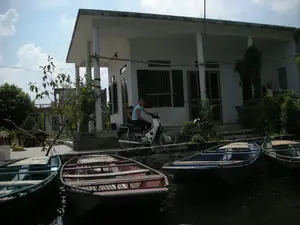 Bối cảnh là một khu vực ven sông hoặc kênh rạch ở một vùng quê, có thể là một điểm du lịch tại Ninh Bình. Trong ảnh có một ngôi nhà một tầng, sơn màu trắng, với mái hiên được chống đỡ bởi các cột tròn. Phía trước nhà, một người phụ nữ mặc áo phông xanh và một người khác đang ngồi trên một chiếc xe máy màu trắng. Dưới mặt nước ở tiền cảnh, nhiều chiếc thuyền nan (ghe) dùng để chở khách du lịch đang neo đậu san sát nhau. Những chiếc thuyền này có các tấm ván gỗ bắc ngang làm ghế ngồi. Xung quanh nhà có cây cối và cây leo. Ở khoảng sân bên trái, có một con vật trông giống chó đang nằm và một nông cụ có thể là máy cày tay. Không có văn bản hay chữ viết nào có thể đọc được trong hình ảnh.。（日本語訳）。背景は、ある田舎の川沿いまたは運河沿いの地域で、ニンビン省の観光地かもしれません。家の前では、青いTシャツを着た女性ともう一人の人が白いバイクに座っています。前景の水面には、観光客を乗せるための多くの竹製のボート（ゲ）が密集して停泊しています。これらのボートには、座席として横に渡された木製の板があります。家の周りには木々やつる植物があります。左側の庭には、犬のような動物が横たわっており、手押し耕運機と思われる農具があります。画像内には読み取れるテキストや文字はありません。