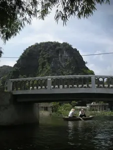 Bức ảnh ghi lại một khung cảnh yên bình trên một con sông ở vùng nông thôn, có khả năng là ở Ninh Bình. Nổi bật trong ảnh là một ngọn núi đá vôi lớn, đặc trưng của khu vực, được cây cối bao phủ. Một cây cầu bê tông thấp bắc ngang qua sông. Dưới gầm cầu, một chiếc thuyền nhỏ (ghe) đang di chuyển, trên thuyền có hai người. Một người trong số họ đội nón lá và đang dùng mái chèo để khua nước. Khung cảnh được nhìn từ một góc thấp, với những cành lá tre rủ xuống ở phía trên, tạo nên một khung hình tự nhiên. Phía xa sau cây cầu là những ngôi nhà và cây cối rậm rạp ven sông.。（日本語訳）。この写真は、田舎の川の穏やかな風景を記録したもので、おそらくニンビンでの一枚です。写真の中で際立っているのは、この地域に特徴的な、木々に覆われた大きな石灰岩の山です。低いコンクリートの橋が川に架かっています。橋の下では、小さなボート（ghe）が移動しており、ボートには2人が乗っています。そのうちの一人はノンラーをかぶり、オールを使って水を漕いでいます。この風景は低いアングルから見られており、上部から垂れ下がる竹の枝葉が自然な額縁を作り出しています。橋の向こうの遠くには、川沿いの家々と鬱蒼とした木々があります。