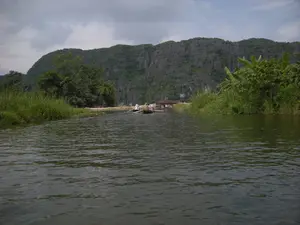 Bức ảnh ghi lại cảnh quan trên một con sông ở khu vực có nhiều núi đá vôi. Dòng sông có màu xanh lục, gợn sóng nhẹ. Ở giữa sông, có một vài chiếc thuyền nan (ghe) đang di chuyển. Một chiếc thuyền có người chèo, có thể là người dân địa phương, đang chở một nhóm du khách đi tham quan. Một trong những du khách đội nón lá. Hai bên bờ sông là thảm thực vật xanh tươi với cỏ lau và cây cối, bao gồm cả những bụi chuối. Phía xa, hậu cảnh là một dãy núi đá vôi hùng vĩ. Có một cây cầu nhỏ bắc qua sông và một tấm biển báo màu xanh dựng gần đó, tuy nhiên nội dung trên biển báo không thể đọc được.。（日本語訳）。写真は、多くの石灰岩の山がある地域の川の風景を記録しています。川は緑色で、穏やかに波立っています。川の中ほどには、数隻の竹製のボート（ゲ）が移動しています。一隻のボートには漕ぎ手がおり、おそらく地元の人で、観光客のグループを乗せて観光しています。観光客の一人はノンラー（菅笠）をかぶっています。川の両岸には、ススキや木々、バナナの茂みを含む青々とした植物が生い茂っています。遠くの背景には、雄大な石灰岩の山脈があります。川を渡る小さな橋と、その近くに立てられた青い看板がありますが、看板の内容は読み取れません。