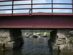 Bức ảnh ghi lại cảnh quan trên một con sông hoặc kênh rạch, được chụp từ vị trí dưới một cây cầu. Cây cầu có kết cấu dầm thép màu đỏ và lan can, được chống đỡ bởi các trụ cầu xây bằng đá. Dưới gầm cầu, một vài chiếc thuyền nhỏ (ghe) đang di chuyển trên mặt nước. Trên thuyền có nhiều người, một số người đội nón lá, đang chèo thuyền đưa khách đi tham quan. Khung cảnh phía sau là những dãy núi đá vôi và cây cối xanh tươi, đặc trưng cho cảnh quan thiên nhiên ở Ninh Bình. Hoạt động chính diễn ra là du ngoạn bằng thuyền trên sông, một hình thức du lịch phổ biến tại khu vực này.。（日本語訳）。写真は、ある川または運河の景観を、橋の下から撮影したものです。橋は赤い鉄骨の梁と欄干の構造で、石造りの橋脚で支えられています。橋の下では、数隻の小舟（サンパン）が水面を移動しています。舟には多くの人が乗っており、何人かはノンラー（菅笠）をかぶり、観光客を乗せて舟を漕いでいます。背景には、ニンビンの自然景観に特徴的な、石灰岩の山々と青々とした木々が広がっています。主なアクティビティは川でのボートツアーで、この地域で人気のある観光形式です。