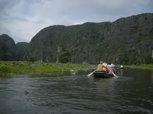 Bối cảnh là một khu vực sông nước với những dãy núi đá vôi hùng vĩ được bao phủ bởi cây cối xanh tươi, có thể là một địa điểm du lịch sinh thái ở Ninh Bình. Trên mặt sông có một vài chiếc thuyền nhỏ (ghe). Chiếc thuyền ở gần nhất đang chở bốn người, trong đó có hai người đang dùng mái chèo để đẩy thuyền đi. Một số người trên thuyền đội nón lá, một loại mũ truyền thống của Việt Nam. Ở phía xa hơn có một chiếc thuyền khác cũng đang di chuyển trên sông. Hoạt động chính là chèo thuyền tham quan, ngắm cảnh. Dọc hai bên bờ sông có nhiều cây sen và các thảm thực vật thủy sinh. Bầu trời có nhiều mây.。（日本語訳）。背景は、青々とした木々に覆われた雄大な石灰岩の山々がある水郷地帯で、おそらくニンビン省のエコツーリズムの観光地でしょう。川面には数隻の小舟（ghe）があります。最も近くにある舟は4人を乗せており、そのうち2人が櫂を使って舟を進めています。舟に乗っている何人かは、ベトナムの伝統的な帽子の一種であるノンラーをかぶっています。さらに遠くには、別の舟も川の上を進んでいます。主なアクティビティは、舟を漕いでの観光や景色を眺めることです。川の両岸に沿って、多くの蓮や水生植物の群生があります。空は雲が多いです。