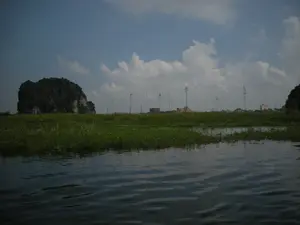 Bức ảnh ghi lại một khung cảnh thiên nhiên tại một khu vực sông nước, có thể là một con sông hoặc vùng đất ngập nước ở Ninh Bình. Không có chữ viết nào có thể nhận dạng được. Tiền cảnh là mặt nước rộng, có những đám thực vật thủy sinh như bèo tây. Ở giữa là một dải đất phủ đầy cỏ xanh. Phía xa, bên trái là một ngọn núi đá vôi lớn, một đặc điểm địa hình quen thuộc của khu vực. Phía sau dải đất, có thể nhìn thấy các tòa nhà của một thị trấn hoặc thành phố. Một vài cột cao, mảnh được dựng lên trên bãi cỏ. Bầu trời trong xanh và có nhiều mây trắng. Toàn bộ khung cảnh cho thấy một chuyến đi trên sông hoặc hồ, ngắm nhìn phong cảnh nơi thiên nhiên giao thoa với khu vực đô thị.。（日本語訳）。写真は、ニンビンの川または湿地である可能性のある、水辺の地域の自然の風景を捉えています。認識できる文字はありません。前景は広い水面で、ホテイアオイのような水生植物の群生があります。中央には、緑の草で覆われた細長い土地があります。遠くの左側には、この地域特有の地形である大きな石灰岩の山があります。細長い土地の後ろには、町または都市の建物が見えます。芝生の上には、いくつかの高くて細い柱が建てられています。空は青く澄んでおり、白い雲が多く浮かんでいます。全体の風景は、川または湖の旅を示しており、自然と都市部が交差する場所の景色を眺めています。