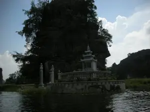 Bức ảnh chụp một khung cảnh ven sông, có khả năng ở khu vực Ninh Bình, nổi bật với một công trình kiến trúc tâm linh. Ở ven bờ sông là một ngôi mộ hoặc am thờ được xây dựng bằng đá trắng. Công trình này có một nhà bia nhỏ với mái cong nhiều tầng ở trung tâm, được bao quanh bởi lan can và có các cột trụ trang trí. Phía sau công trình là một vách núi đá vôi lớn, sẫm màu và hùng vĩ, được bao phủ bởi cây cối xanh tươi. Dưới chân núi và ven bờ là thảm cỏ và cây thủy sinh. Mặt nước sông ở phía trước khá tối, phản chiếu bóng của ngọn núi. Bầu trời có mây trắng trên nền xanh. Không có người hay hoạt động nào đang diễn ra, tạo nên một không gian yên tĩnh và trang nghiêm. Không có chữ viết nào có thể nhận dạng được trên công trình hay bất kỳ vật thể nào khác.。（日本語訳）。写真は川沿いの風景を撮影したもので、おそらくニンビン地方のものであり、精神的な建築物が際立っています。川岸には、白い石で建てられた墓または祠があります。この建造物は、中央に多層の反り屋根を持つ小さな石碑の家があり、手すりで囲まれ、装飾的な柱があります。建造物の後ろには、緑豊かな木々に覆われた、大きくて暗い色の雄大な石灰岩の崖があります。山の麓と岸辺には、草や水生植物が広がっています。手前の川の水面はかなり暗く、山の影を映しています。空は青い背景に白い雲が浮かんでいます。人や活動は見られず、静かで荘厳な空間を作り出しています。建造物や他のどの物体にも、識別できる文字はありません。