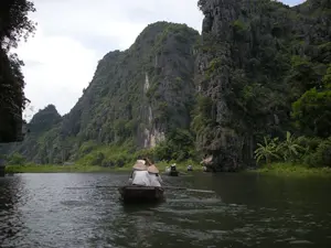 Bức ảnh chụp cảnh du ngoạn trên một con sông hoặc kênh nước, bao quanh bởi những dãy núi đá vôi hùng vĩ. Đây là một khung cảnh đặc trưng của vùng Ninh Bình. Hoạt động chính là tham quan bằng thuyền. Ở tiền cảnh, một chiếc thuyền nan nhỏ đang được một người chèo, chở hai hành khách. Cả hai hành khách đều đội nón lá truyền thống của Việt Nam. Phía xa có thể thấy vài chiếc thuyền tương tự cũng đang chở khách. Phong cảnh hai bên bờ sông là những vách núi đá cao, được cây cối xanh tươi bao phủ. Dưới mặt nước có thể thấy gợn sóng do mái chèo tạo ra. Không có văn bản, chữ viết hay biển hiệu nào trong ảnh.。（日本語訳）。雄大な石灰岩の山々に囲まれた、川または水路での遊覧風景を撮影した写真です。これはニンビン地方の典型的な風景です。主なアクティビティはボートでの観光です。前景には、一人の船頭が漕ぐ小さな竹のボートが写っており、2人の乗客を乗せています。乗客は2人ともベトナムの伝統的なノンラー（菅笠）をかぶっています。遠くには、同じように客を乗せている数隻のボートが見えます。川の両岸の風景は、青々とした木々に覆われた高い岩壁です。水面には、オールによってできた波紋が見えます。写真にはテキスト、文字、看板はありません。