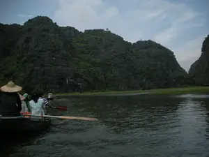 Đây là một cảnh quan sông nước tại một khu du lịch, có thể là ở Ninh Bình, với những dãy núi đá vôi đặc trưng. Hoạt động chính là du ngoạn bằng thuyền. Trong ảnh, có một chiếc thuyền nhỏ (ghe) đang chở một nhóm du khách trên sông. Một người trong nhóm đang chèo thuyền bằng mái chèo gỗ. Một người khác ngồi ở phía sau thuyền đội nón lá. Phía trước có một chiếc thuyền khác cũng đang di chuyển. Xung quanh là những ngọn núi đá vôi cao, phủ đầy cây xanh. Trên đỉnh một ngọn núi ở phía xa có một công trình kiến trúc nhỏ, có thể là một ngôi chùa hoặc vọng lâu. Bầu trời u ám và có mây.。（日本語訳）。これは、特徴的な石灰岩の山々がある、ある観光地の水辺の風景で、おそらくニンビンでしょう。主なアクティビティはボートでの遊覧です。写真の中では、一艘の小さなボート（ghe）が川で観光客のグループを乗せています。グループの一人が木製のオールでボートを漕いでいます。もう一人はボートの後ろに座り、ノンラーをかぶっています。前方には、別のボートも移動しています。周囲は、緑に覆われた高い石灰岩の山々です。遠くの山の頂上には、小さな建築物があり、それは寺院か望楼かもしれません。空はどんよりと曇っています。
