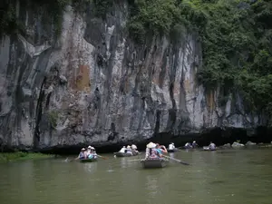 Bức ảnh ghi lại cảnh một đoàn du khách đang đi thuyền tham quan trên một dòng sông. Địa điểm là một khu vực có cảnh quan núi đá vôi hùng vĩ, có thể là một khu du lịch sinh thái ở Ninh Bình. Có nhiều chiếc thuyền nan (ghe) nhỏ đang di chuyển trên mặt nước màu xanh đục. Trên mỗi thuyền là các du khách và một người chèo thuyền, một số người đội nón lá. Các chiếc thuyền đang đi dọc theo chân một vách núi đá lớn, sừng sững với cây cối mọc xanh tốt ở phía trên. Hoạt động chính là du ngoạn, ngắm cảnh thiên nhiên. Không có bất kỳ văn bản, biển hiệu hay chữ viết nào trong ảnh.。（日本語訳）。写真は、一団の観光客が川で船に乗って観光している光景を捉えています。場所は雄大な石灰岩の山の景観がある地域で、おそらくニンビン省のエコツーリズム地区でしょう。濁った緑色の水面の上を、多くの小さな竹製のボート（ゲ）が移動しています。各ボートには観光客と一人の船頭が乗っており、何人かはノンラー（菅笠）をかぶっています。ボートは、上部に青々と茂る木々が生い茂る、そびえ立つ大きな岩壁の麓に沿って進んでいます。主なアクティビティは遊覧と自然景観の鑑賞です。写真の中には、いかなるテキスト、看板、文字もありません。