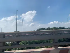 Bức ảnh được chụp từ một phương tiện đang di chuyển, nhìn ra một cây cầu vượt hoặc một đoạn đường trên cao. Khung cảnh cho thấy một cây cầu bê tông với lan can và dải phân cách được sơn sọc đỏ và trắng. Dọc theo cây cầu là nhiều cột đèn đường cao. Phía sau cây cầu là một hàng cây xanh rậm rạp, bao gồm cả cây cọ. Bầu trời trong xanh với những đám mây trắng, cho thấy thời tiết nắng đẹp. Không có văn bản hay biển báo nào có thể nhìn thấy rõ. Không có hoạt động cụ thể nào của con người được quan sát, chỉ có cảnh quan giao thông đường bộ.。（日本語訳）。移動中の乗り物から撮影された写真で、陸橋または高架道路が見えます。橋に沿って、背の高い街灯がいくつも立っています。橋の後ろには、ヤシの木を含む緑豊かな木々が立ち並んでいます。空は青く澄み渡り、白い雲が浮かんでおり、晴天であることを示しています。特定の人間の活動は見られず、道路交通の風景だけが観察されます。