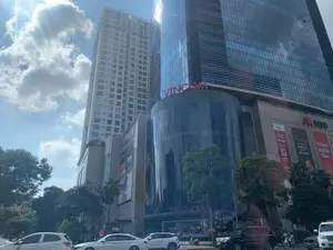 Bức ảnh chụp một cảnh quan đô thị hiện đại tại Hà Nội, với các tòa nhà chọc trời dưới bầu trời xanh và mây trắng. Đây là một khu vực thương mại và văn phòng sầm uất.。**Chữ viết được ghi lại:**。*   Trên tòa nhà chính bằng kính, có dòng chữ "VINCOM" và "VINCOM CENTER" màu đỏ.。*   Một tòa nhà liền kề có logo của ngân hàng "MSB" và biển hiệu của trung tâm thể hình "ELITE FITNESS".。*   Các biểu ngữ quảng cáo lớn có thể đọc được bao gồm: "TNL", "CƠ HỘI THUÊ VĂN PHÒNG", "TNR TOWER", "TẶNG ĐẾN 05 THÁNG TIỀN THUÊ"; "Thẻ mDigi", "LINH HOẠT HOÀN TIỀN", "TỰ DO SỐNG CHẤT"; và "CÙNG VƯƠN TẦM".。*   Có thể nhận ra một phần tên thương hiệu "PHUC LONG" và "HIGHLANDS COFFEE" ở tầng trệt.。**Bối cảnh và hoạt động:**。*   **Địa điểm:** Một con đường lớn phía trước trung tâm thương mại Vincom và tòa nhà văn phòng ở Hà Nội.。*   **Hoạt động:** Dưới lòng đường, nhiều ô tô và xe máy đang tham gia giao thông, thể hiện nhịp sống đô thị nhộn nhịp.。*   **Vật thể:** Các tòa nhà cao tầng hiện đại, ô tô, xe máy, cây xanh đô thị và các biển quảng cáo.。（日本語訳）。写真は、青い空と白い雲の下に高層ビルがそびえ立つ、ハノイの現代的な都市景観を写したものです。ここは賑やかな商業・オフィスエリアです。**記録された文字：**。*   ガラス張りの主要な建物には、赤い文字で「VINCOM」と「VINCOM CENTER」と書かれています。*   隣接する建物には、「MSB」銀行のロゴと「ELITE FITNESS」フィットネスセンターの看板があります。*   読み取れる大きな広告バナーには、「TNL」、「CƠ HỘI THUÊ VĂN PHÒNG」、「TNR TOWER」、「TẶNG ĐẾN 05 THÁNG TIỀN THUÊ」；「Thẻ mDigi」、「LINH HOẠT HOÀN TIỀN」、「TỰ DO SỐNG CHẤT」；そして「CÙNG VƯƠN TẦM」が含まれます。*   1階には、「PHUC LONG」と「HIGHLANDS COFFEE」のブランド名の一部が認識できます。**背景と活動：**。*   **場所：** ハノイのビンコム商業センターとオフィスビルの前にある大通り。*   **活動：** 路上では、多くの車やバイクが往来しており、賑やかな都市の生活リズムを表しています。*   **物体：** 現代的な高層ビル、車、バイク、都市の緑樹、そして広告看板。