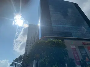 Bức ảnh chụp một tòa nhà chọc trời hiện đại với mặt tiền bằng kính, được chụp từ góc nhìn thấp hướng lên vào một ngày nắng đẹp. Bầu trời trong xanh với vài gợn mây trắng, và mặt trời tỏa sáng rực rỡ. Phía trước tòa nhà có một cây xanh lớn, tán lá che một phần chân tòa nhà. Đây là một khu phức hợp thương mại và văn phòng hiện đại.。Các chữ viết trên biển hiệu và biểu ngữ quảng cáo có thể nhận dạng được bao gồm:。- Logo "VINCOM" màu đỏ.。- Một biểu ngữ quảng cáo có nội dung: "#TNL", "CƠ HỘI THUÊ VĂN PHÒNG", "TNR TOWER", "TẶNG ĐẾN 05 THÁNG TIỀN THUÊ", "3 NĂM PHÍ GỬI XE".。- Một biểu ngữ khác bên cạnh có các từ: "LINH HOẠT", "HOÀN TIỀN", "TỰ DO", "SỐNG CHẤT".。Khung cảnh cho thấy đây là một khu đô thị sầm uất, cụ thể là bên ngoài tòa nhà TNR Tower, nơi có trung tâm thương mại Vincom, với các hoạt động quảng cáo cho thuê văn phòng.。（日本語訳）。写真は、晴れた日に下から見上げる角度で撮影された、ガラス張りのファサードを持つ近代的な超高層ビルを写しています。空は青く澄み渡り、白い雲がいくつか浮かんでおり、太陽がまぶしく輝いています。ビルの前には大きな緑の木があり、その葉がビルの一階部分を一部覆っています。これは近代的な商業およびオフィス複合施設です。看板や広告バナーに書かれている識別可能な文字には、以下のものが含まれます：。- 赤い「VINCOM」のロゴ。- 「#TNL」、「オフィス賃貸の機会」、「TNR TOWER」、「最大5ヶ月分の賃料をプレゼント」、「3年間の駐車料金」という内容の広告バナー。- 隣にある別のバナーには、「柔軟」、「返金」、「自由」、「質の高い生活」という言葉があります。この光景は、ここが賑やかな都市部、具体的にはオフィス賃貸の広告活動が行われている、VincomショッピングセンターがあるTNRタワーの外であることを示しています。