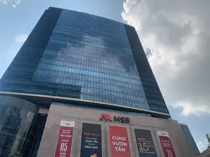 Bức ảnh chụp góc nhìn từ dưới lên của một tòa nhà chọc trời hiện đại với mặt tiền bằng kính màu xanh, phản chiếu bầu trời có mây. Đây là một tòa nhà văn phòng thương mại.。Phần đế của tòa nhà có gắn logo lớn của 'MSB' và một loạt các áp phích quảng cáo. Toàn bộ văn bản có thể đọc được bao gồm:。- Logo: MSB。- Áp phích 1 (bên trái): '#TNL', 'CƠ HỘI THUÊ VĂN PHÒNG', 'TNR TOWER', 'TẶNG ĐẾN 05 THÁNG TIỀN THUÊ'.。- Áp phích 2: 'Thẻ mDigi', 'LINH HOẠT HOÀN TIỀN', 'TỰ DO SỐNG CHẤT'.。- Áp phích 3: 'CÙNG VƯƠN TẦM'.。- Áp phích 4: 'M-POWER', 'CẤP TÍN CHẤP ĐẾN 15 TỶ ĐỒNG', 'dành cho Doanh nghiệp'.。- Áp phích 5 (bên phải): '#TNI', 'ĐƠN VỊ PHÁT TRIỂN KHU CÔNG NGHIỆP', 'HÀNG ĐẦU VIỆT NAM', và số '20'.。Cảnh vật cho thấy đây là một tòa nhà văn phòng hiện đại, nơi diễn ra các hoạt động thương mại và cho thuê văn phòng, được quảng bá qua các tấm áp phích.。（日本語訳）。青いガラスのファサードが曇り空を反射している、現代的な高層ビルを下から見上げた写真です。これは商業オフィスビルです。ビルの基部には「MSB」の大きなロゴと一連の広告ポスターが掲示されています。読み取れるすべてのテキストは以下の通りです。- ロゴ：MSB。- ポスター1（左側）：「#TNL」、「オフィス賃貸の機会」、「TNR TOWER」、「最大5ヶ月分の賃料をプレゼント」。- ポスター2：「mDigiカード」、「柔軟なキャッシュバック」、「自由に質の高い生活を」。- ポスター3：「共に高みへ」。- ポスター4：「M-POWER」、「最大150億ドンの無担保融資」、「企業向け」。- ポスター5（右側）：「#TNI」、「工業団地開発ユニット」、「ベトナムのトップ」、そして数字の「20」。この光景は、ここが現代的なオフィスビルであり、商業活動やオフィス賃貸が行われ、それらがポスターを通じて宣伝されていることを示しています。