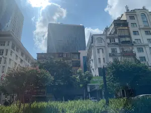 写真は、晴れた美しい日、青空に雲が浮かぶハノイの街並みを撮影したものです。新旧の建築が混在しています。後方にはガラス張りの高層ビルがあり、その隣にある古い様式の多層階建ての建物と対照をなしています。古い建物にはバルコニー、エアコンの室外機、屋上には通信アンテナがあります。前景には緑の木々と、鮮やかなピンク色の花を咲かせた木々があります。左側にはバイクに乗っている男性がいます。多くの看板が見え、そのうちいくつかは読み取ることができます。緑色の看板には「BỆNH VIỆN BA...」と住所「52 Nguyễn Chí Thanh」と書かれています。他のレストランの看板には、「Nhà Hàng Hải Sản Biển Xanh」、「PHO NOI」、そして「... CỦA PHÚ SƯỜN」という看板の一部が含まれています。この光景は、大都市に特徴的な、にぎやかな街角の一場面を表しています