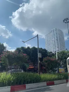 Bức ảnh chụp một con đường tại Hà Nội vào một ngày nắng đẹp, bầu trời trong xanh với những đám mây trắng. Ở hậu cảnh là một tòa nhà cao tầng hiện đại màu trắng với nhiều cửa sổ. Dọc hai bên đường và trên dải phân cách là cây xanh, trong đó có những cây bằng lăng đang nở hoa màu hồng rực rỡ. Dải phân cách ở giữa đường được trồng cỏ và có bó vỉa sơn màu đỏ và trắng. Phía bên kia đường là một dãy các cửa hàng san sát nhau. Có thể đọc được một số biển hiệu quảng cáo: "NGUYÊN NHÀ MOBILE" với các dịch vụ "MUA BÁN - SỬA CHỮA", "LẤY NGAY"; cửa hàng "DIỆP HÀ Mobile" chuyên "BÁN ĐIỆN THOẠI", "SỬA CHỮA ÉP KÍNH"; và một cửa hàng có biển hiệu "Fruits". Ngoài ra còn có các biển hiệu khác ghi "TẶNG", "BẢO N". Các vật thể khác trong ảnh bao gồm cột đèn đường, camera giám sát giao thông, và một phần của một chiếc xe buýt hoặc xe tải ở rìa phải bức ảnh.。（日本語訳）。写真はハノイのある晴れた美しい日に撮影されたもので、空は青く澄み渡り、白い雲が浮かんでいる。背景には、多くの窓がある近代的な白い高層ビルがある。道路の両側と中央分離帯には緑の木々があり、その中には鮮やかなピンク色の花を咲かせているサルスベリの木もある。道路の中央分離帯には草が植えられ、縁石は赤と白に塗装されている。道路の向こう側には、店がずらりと並んでいる。いくつかの広告看板が読み取れる：「NGUYÊN NHÀ MOBILE」は「売買 - 修理」「即日対応」のサービスを提供。「DIỆP HÀ Mobile」店は「携帯電話販売」「修理・ガラス圧着」を専門としている。そして「Fruits」という看板の店もある。その他にも、「TẶNG（贈呈）」、「BẢO N」と書かれた看板もある。写真の中のその他の物体には、街灯、交通監視カメラ、そして写真の右端にあるバスまたはトラックの一部が含まれる。