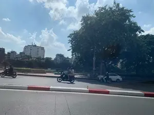 Đây là một cảnh đường phố ở Hà Nội vào một ngày nắng đẹp. Trên con đường nhựa rộng rãi, có nhiều người đang tham gia giao thông bằng xe máy. Có thể thấy một vài chiếc xe máy và một chiếc ô tô màu trắng. Một số người đi xe máy một mình, trong khi một chiếc xe khác chở hai người. Tất cả mọi người đều đội mũ bảo hiểm. Bên phải đường là một cây cổ thụ lớn với tán lá xanh rậm rạp. Phía xa, bên kia con đường là các tòa nhà cao tầng và dường như có một con sông hoặc kênh chạy dọc theo. Vỉa hè được sơn vạch đỏ và trắng. Bầu trời trong xanh với những đám mây trắng. Trên một tòa nhà ở phía xa có một biển hiệu chữ nhưng khó đọc rõ. Một biểu ngữ cũng được treo trên cột đèn nhưng không thể đọc được nội dung.。（日本語訳）。これは晴れた日のハノイの街の風景です。広々としたアスファルトの道路では、多くの人々がバイクで交通に参加しています。数台のバイクと一台の白い車が見えます。一人でバイクに乗っている人もいれば、二人乗りをしているバイクもあります。全員がヘルメットを着用しています。道の右側には、青々とした葉が茂る大きな古い木があります。遠く、道の向こう側には高層ビルがあり、それに沿って川か運河が流れているようです。歩道は赤と白の線で塗られています。空は青く澄み渡り、白い雲が浮かんでいます。遠くの建物の一つには文字の看板がありますが、はっきりと読み取るのは困難です。街灯の柱にも横断幕が掲げられていますが、その内容は読み取れません。