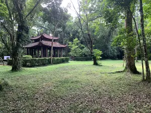 緑豊かな森の中に建つ、伝統的な様式の木造の東屋。手前は芝生の広場になっている。遠方に看板らしきものがあるが、文字は