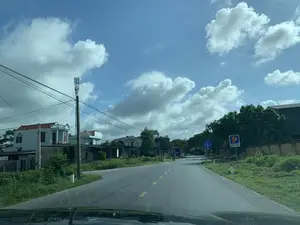 Bức ảnh chụp quang cảnh một con đường nông thôn ở khu vực Nga Sơn, nhìn từ bên trong một chiếc ô tô. Bầu trời trong xanh với nhiều đám mây trắng. Con đường được trải nhựa, có vạch kẻ đường màu vàng ở giữa.。Hai bên đường là những ngôi nhà dân, một số nhà được xây dựng theo kiểu hiện đại, xen kẽ với cây cối và thảm thực vật xanh tươi. Có nhiều cột điện và dây điện dọc theo con đường. Một cột thu phát sóng di động (trạm BTS) cao được dựng ở bên trái.。Bên phải đường có một số biển báo. Một biển báo chỉ dẫn giao thông màu xanh lam ghi:。- Hướng đi thẳng: NGA SƠN。- Hướng rẽ phải: NGÃ THỦY。Gần đó là một biển báo quảng cáo của Petrolimex với logo chữ "P" và dòng chữ "CÁCH 250 M", cho biết có một trạm xăng ở phía trước 250 mét.。Khung cảnh yên tĩnh, không có nhiều hoạt động. Ở phía xa có một người đang đi xe máy trên đường.。（日本語訳）。ガーソン地区の田舎道の光景を車内から撮影した写真です。空は青く澄んでおり、白い雲がたくさん浮かんでいます。道路は舗装されており、中央には黄色い車線が引かれています。道路の両側には民家があり、いくつかの家は現代的な様式で建てられ、木々や青々とした草木と入り混じっています。道路に沿ってたくさんの電柱と電線があります。左側には、一本の高い携帯電話基地局（BTS局）が建てられています。道路の右側には、いくつかの標識があります。一つの青い交通案内標識には、次のように書かれています。- 直進方向：ガーソン。- 右折方向：ガー・トゥイ。その近くには、「P」の文字のロゴと「250M先」という文字が書かれたペトロリメックス（Petrolimex）の広告看板があり、250メートル先にガソリンスタンドがあることを示しています。静かな光景で、あまり動きがありません。遠くには、道路をバイクで走っている人が一人います。