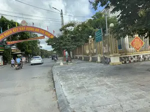 Bức ảnh chụp một con đường tại một khu đô thị ở Việt Nam, có thể là huyện Nga Sơn. Khung cảnh cho thấy hoạt động giao thông và sinh hoạt thường ngày. Trên đường có một chiếc ô tô màu trắng và một người đang lái xe máy. Ở lề đường bên trái, có hai người đang đi bộ. Vỉa hè được lát gạch đá và hai bên đường có nhiều cây xanh, trong đó có một cây nhãn. Nổi bật trong ảnh là một cổng chào lớn màu đỏ bắc ngang đường với dòng chữ vàng: '...ĐOÀN KẾT XÂY DỰNG ĐÔ THỊ VĂN MINH'. Bên phải là một tòa nhà có hàng rào kim loại màu vàng, trên đó có các biển báo. Một biển chỉ dẫn màu xanh lá cây ghi: 'NGÂN HÀNG CHÍNH SÁCH XÃ HỘI HUYỆN NGA SƠN' và 'Cách 200m'. Một biển báo khác hình thoi màu vàng có nội dung: 'KHOA HỌC CÔNG NGHỆ, ĐỔI MỚI SÁNG TẠO VÀ CHUYỂN ĐỔI SỐ LÀ ĐỘNG LỰC CHÍNH CỦA TĂNG TRƯỞNG KINH TẾ'. Ở phía xa bên trái có các cửa hàng với các biển hiệu như 'BUN' và 'PHÚC LỘC'.。（日本語訳）。写真はベトナムのある都市部の道路を撮影したもので、おそらくガーソン県だと思われます。この光景は、日常の交通と生活の様子を示しています。道路には白い自動車一台と、バイクを運転している人が一人います。左側の路肩には、歩いている人が二人います。歩道は石タイルで舗装されており、道の両側には多くの緑の木があり、その中にはリュウガンの木が一本あります。写真の中で目立っているのは、道をまたぐように建てられた大きな赤い歓迎ゲートで、そこには黄色い文字で「...団結して文明的な都市を建設する」と書かれています。右側には黄色い金属のフェンスがある建物があり、その上には看板がいくつかあります。緑色の案内標識の一つには、「ガーソン県社会政策銀行」そして「200m先」と書かれています。もう一つの黄色い菱形の看板には、「科学技術、イノベーション、そしてデジタルトランスフォーメーションは経済成長の主要な原動力である」という内容が書かれています。遠くの左側には、「BUN」や「PHÚC LỘC」といった看板を掲げた店があります。