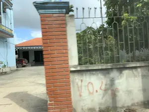 Hình ảnh ghi lại một khung cảnh ven đường tại một khu dân cư, có vẻ như được chụp từ một phương tiện đang di chuyển. Ở phía trước, một cột trụ xây bằng gạch đỏ và một bức tường bê tông bị nhòe do chuyển động. Trên tường bê tông có các ký tự được vẽ bằng sơn màu đỏ nhưng không thể đọc rõ. Phía sau tường là một hàng rào sắt màu xanh lá cây và cây cối. Xa hơn là một ngôi nhà mái ngói đỏ với khoảng sân rộng, nơi có một chiếc ô tô màu đỏ đang đỗ. Bên cạnh, một phần của một ngôi nhà khác sơn màu xanh nhạt cũng có thể được nhìn thấy. Bầu trời có mây.。（日本語訳）。画像は、ある住宅地の道端の風景を記録したもので、移動中の乗り物から撮影されたようです。手前には、赤レンガで造られた柱とコンクリートの壁が、動きによってぼやけています。壁の後ろには、緑色の鉄柵と木々があります。さらに奥には、赤い瓦屋根の家があり、広い庭には赤い車が一台駐車しています。その隣には、水色に塗られた別の家の一部も見えます。空は曇っています。