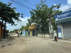 Bản ghi chép văn bản:。- Biển hiệu màu xanh dương bên phải: "Amozo SƠN & CHỐNG THẤM. ĐỊA CHỈ: XÓM 1 - NGA THỦY - NGA SƠN - THANH HÓA. NPP: HƯNG CƯỜNG. UY TÍN LÀM NÊN TH...". Phía dưới là một biển hiệu nhỏ hơn: "Amozo. TƯ VẤN CUNG CẤP VẬT TƯ, CHỐNG THẤM SIKA, SÀN MÁI, TƯỜNG ĐỨNG. ĐỊA CHỈ: NGA THỦY NGA SƠN - THANH HÓA."。- Biển hiệu màu vàng ở giữa: "THẾ GIỚI TƯƠNG LAI 68 mobile. SỬA CHỮA - BẢO HÀNH ĐIỆN THOẠI CHÍNH HÃNG. BÁN PHỤ KIỆN CHÍNH HÃNG. ĐT: 0968 985 625."。Mô tả cảnh quan và hoạt động:。- Loại địa điểm: Một con đường đất tại một ngôi làng hoặc thị trấn nhỏ ở vùng nông thôn Việt Nam. Hai bên đường là nhà dân và các cửa hàng, bao gồm một cửa hàng bán sơn và vật liệu xây dựng, và một cửa hàng sửa chữa điện thoại di động.。- Hoạt động cụ thể: Cảnh sinh hoạt đời thường. Một người phụ nữ đội nón lá đang đi bộ bên lề đường, tay xách túi đồ, có vẻ như đang trên đường đi chợ hoặc từ chợ về. Ở phía xa hơn, một người khác cũng đội nón lá đang đi xe đạp. Có người đang đi xe máy ở phía sau.。- Các vật thể được ghi lại: Nhà cửa, cửa hàng với các biển hiệu quảng cáo. Người dân trong trang phục thường ngày, đặc biệt là hình ảnh người đội nón lá. Phương tiện di chuyển gồm xe đạp và xe máy. Dọc đường có cây xanh và các cột điện với hệ thống dây điện chằng chịt.。（日本語訳）。テキストの書き起こし：。- 右側の青い看板：「Amozo 塗料＆防水。住所：タインホア省ガソン県ガトゥイ社第1集落。販売代理店：フンクオン。信頼が創り出す…」。その下にはより小さな看板：「Amozo。資材のコンサルティング・供給、シカ防水、屋上床、垂直壁。住所：タインホア省ガソン県ガトゥイ社。」。- 中央の黄色い看板：「THE GIOI TUONG LAI 68 mobile。純正携帯電話の修理・保証。純正アクセサリー販売。電話番号：0968 985 625。」。風景と活動の描写：。- 場所の種類：ベトナムの田舎にある村または小さな町の未舗装の道。道の両側には民家や店があり、塗料・建材店や携帯電話修理店などがある。- 具体的な活動：日常生活の光景。ノンラーをかぶった女性が道端を歩いており、手には荷物袋を提げている。市場へ行く途中か、市場からの帰り道のようだ。さらに遠くには、同じくノンラーをかぶった別の人が自転車に乗っている。後方にはバイクに乗っている人がいる。- 記録された物体：家、広告看板のある店。普段着の人々、特にノンラーをかぶった人々の姿。移動手段には自転車やバイクが含まれる。道沿いには緑の木々と、複雑に絡み合った電線のある電柱がある。