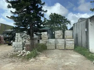 Đây là một khu vực trông giống như một bãi vật liệu xây dựng hoặc sân của một nhà máy. Có rất nhiều chồng gạch được xếp trên các pallet gỗ và được bọc trong nilon. Có cả gạch màu sáng (gạch không nung) và gạch nung màu đỏ. Ở phía sau bên trái, một chiếc xe tải lớn màu đen đang đỗ. Trên kính chắn gió của xe có chữ 'SM'. Bên phải là một tòa nhà có tường bê tông đã cũ. Phía sau có các công trình lợp mái tôn màu xanh và nhiều cây cối. Một cây tùng lớn đứng ở phía trước. Mặt đất là đất bụi, có cỏ dại và gạch vỡ vương vãi. Bầu trời xanh và có mây. Không có người hay hoạt động nào đang diễn ra, cho thấy đây là một khu vực lưu trữ.。（日本語訳）。木製パレットの上に積まれ、ナイロンで包まれたレンガの山がたくさんあります。明るい色のレンガ（非焼成レンガ）と赤色の焼成レンガの両方があります。左奥には、大きな黒いトラックが駐車しています。その車のフロントガラスには「SM」という文字があります。右側には、古いコンクリートの壁の建物があります。その後ろには、青いトタン屋根の建物や多くの木々があります。手前には大きな松の木が立っています。地面は土埃で、雑草や割れたレンガが散らばっています。空は青く、雲があります。人や活動の様子はなく、ここが保管エリアであることを示しています。