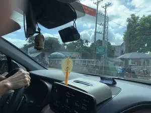 Bức ảnh được chụp từ góc nhìn bên trong một chiếc ô tô, nhìn ra ngoài qua kính chắn gió. Khung cảnh bên ngoài là một con đường ở Việt Nam, có thể là khu vực nông thôn hoặc thị trấn. Dọc hai bên đường có nhà cửa, cây xanh và nhiều cột điện. Có một biển báo màu xanh lá cây ghi rõ 'ĐIỂM GIAO DỊCH XÃ NGA TÂN'. Ngoài ra, có một băng rôn tuyên truyền màu đỏ treo trên cột điện với dòng chữ có thể đọc được một phần là '... VỚI CHỦ ĐỀ: ... TÚY'. Có một vài người đang di chuyển bằng xe máy và các quầy hàng ven đường được che bằng ô lớn, cho thấy có hoạt động buôn bán. Một cây cầu bê tông nhỏ cũng có thể được nhìn thấy. Bên trong xe, tay của người lái đang đặt trên vô lăng. Trên bảng điều khiển là một màn hình hiển thị thông tin như thời gian 8:26 sáng, ngày 08.07, và nhiệt độ 23.5°C. Ngoài ra còn có một hộp khăn giấy và các vật trang trí như sáp thơm treo trên gương chiếu hậu.。（日本語訳）。写真は一台の車の中から、フロントガラスを通して外を見た視点で撮られています。外の風景はベトナムのある道路で、田舎の地域か町かもしれません。道路の両脇には家々、緑の木々、そして多くの電柱があります。「ĐIỂM GIAO DỊCH XÃ NGA TÂN」とはっきりと書かれた緑色の看板があります。さらに、電柱には赤色の宣伝用の横断幕が掛けられており、その文字は「... VỚI CHỦ ĐỀ: ... TÚY」と部分的に読むことができます。バイクで移動している数人の人々と、大きなパラソルで覆われた道端の屋台があり、商売活動が行われていることを示しています。小さなコンクリートの橋も見えます。車内では、運転手の手がハンドルに置かれています。ダッシュボードの上には、午前8時26分、7月8日、そして気温23.5℃といった情報を表示する画面があります。さらに、ティッシュペーパーの箱や、バックミラーに吊るされた芳香剤のような装飾品もあります。