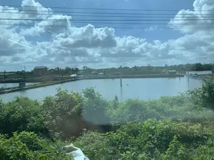 この写真は、農村地域の水産養殖場を描写しています。風景は主に大きな池や沼であり、エビや魚の養殖場である可能性があります。これらの池は土手で互いに隔てられており、養殖を補助するための水車システム（曝気装置）が設置されています。池の周りには木々が鬱蒼と茂っています。遠くにはいくつかの民家が見え、そのうちの一軒は建設中のようです。空は青く澄んでおり、白い雲が多く浮かび、上空には電線が横切っています。写真の中には、識別できる文字や看板はありません