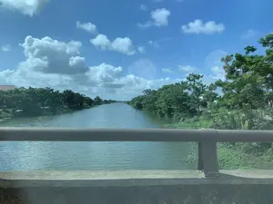 Bức ảnh chụp khung cảnh một con sông nhìn từ trên một cây cầu. Ở tiền cảnh là lan can cầu bằng bê tông và một thanh chắn kim loại màu nâu. Dòng sông rộng, mặt nước phẳng lặng. Hai bên bờ sông là những hàng cây xanh tươi tốt rậm rạp. Phía bờ bên trái, có thể thấy một phần mái nhà ngói đỏ ẩn sau lùm cây. Bầu trời trong xanh, có nhiều mây trắng. Toàn cảnh cho thấy một khung cảnh làng quê yên bình và đặc trưng.。（日本語訳）。橋の上から見た川の風景を写した写真です。前景には、コンクリート製の橋の欄干と茶色の金属製の手すりがあります。川は広く、水面は穏やかです。川の両岸には、青々と生い茂る木々が並んでいます。左岸には、木々の茂みの後ろに隠れた赤い瓦屋根の一部が見えます。空は青く澄んでおり、白い雲が多く浮かんでいます。全景は、平穏で特徴的な田舎の風景を示しています。