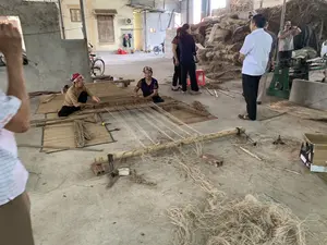Bối cảnh là một xưởng sản xuất thủ công, có thể là một cơ sở trong làng nghề dệt chiếu. Hai người phụ nữ đang ngồi trên sàn xi măng, sử dụng một khung cửi thủ công bằng gỗ và tre để dệt chiếu. Họ đội khăn trên đầu. Xung quanh họ là các tấm chiếu đang được dệt và các bó sợi cói (nguyên liệu dệt). Một nhóm người, có vẻ là khách tham quan, đang đứng xem quá trình làm việc. Trong nhóm này, có một người đàn ông mặc áo sơ mi trắng và một người đàn ông khác đang dùng điện thoại để quay phim hoặc chụp ảnh. Ở phía sau, có những đống sợi cói khô rất lớn, một vài chiếc xe đạp và một số máy móc. Không có chữ viết hay biển hiệu nào có thể đọc rõ.。（日本語訳）。背景は手工業の作業場で、ござ織りの伝統工芸村の施設かもしれません。二人の女性がセメントの床に座り、木と竹でできた手織り機を使ってござを織っています。彼女たちは頭にスカーフを巻いています。彼女たちの周りには、織りかけのござや、い草の束（織物の原料）があります。見物客と思われる一団が、作業の様子を立って見ています。このグループの中には、白いシャツを着た男性と、スマートフォンで動画か写真を撮影している別の男性がいます。後方には、乾燥したい草の大きな山、数台の自転車、そしていくつかの機械があります。はっきりと読み取れる文字や看板はありません。