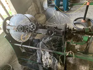 Bối cảnh là một xưởng sản xuất thủ công, có thể là một xưởng se sợi cói. Sàn nhà bê tông bừa bộn với các sợi cói khô. Ở trung tâm bức ảnh là một cỗ máy đang hoạt động. Cỗ máy này có cấu trúc phức tạp với nhiều bánh răng kim loại, bánh đà và dây curoa, được dùng để bện các sợi cói khô thành dây thừng hoặc sợi lớn hơn. Có một cuộn dây cói thành phẩm nằm ở phía sau. Một người mặc quần dài màu xanh lá cây đang đứng gần đó, có lẽ đang vận hành hoặc giám sát máy. Rải rác trên sàn là rất nhiều sợi cói. Trên một bộ phận tròn, lớn, màu đen của máy có dòng chữ màu vàng, có thể đọc được một phần là 'SOLID... PREMIUM...'. Có những tảng đá lớn được đặt gần máy, có thể để giữ cho máy ổn định. Khung cảnh cho thấy một công đoạn trong quá trình sản xuất các sản phẩm từ cói, một nghề thủ công truyền thống.。（日本語訳）。背景は手工芸の作業場、おそらく藺草の糸を撚る作業場です。コンクリートの床は、乾燥した藺草の繊維で散らかっています。写真の中央には、稼働中の機械があります。この機械は、多くの金属製の歯車、フライホイール、ベルトコンベアを備えた複雑な構造をしており、乾燥した藺草の繊維を編んでロープやより太い糸にするために使われます。後ろには、完成した藺草のロープの巻物があります。緑色のズボンを履いた人が近くに立っており、おそらく機械を操作または監督しています。床には非常に多くの藺草の繊維が散らばっています。機械の黒くて大きくて丸い部品の上には黄色い文字があり、「SOLID... PREMIUM...」と一部読み取れます。機械の近くには大きな石が置かれており、おそらく機械を安定させるためのものです。この光景は、伝統的な手工芸である藺草製品の製造過程の一工程を示しています。