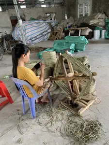 Bối cảnh là một nhà xưởng hoặc nhà kho sản xuất thủ công. Một người phụ nữ mặc áo phông màu vàng đang ngồi trên ghế nhựa màu xanh, tập trung vào công việc se và quấn dây thừng làm từ sợi thực vật, có thể là sợi cói. Cô ấy đang sử dụng một chiếc guồng quay bằng gỗ để cuộn các sợi dây lại với nhau. Xung quanh cô có nhiều cuộn dây thành phẩm đã được buộc gọn gàng và xếp chồng lên nhau. Trên sàn nhà bê tông có nhiều sợi vụn vương vãi. Trong hậu cảnh của nhà xưởng, có thể thấy một chiếc xe đạp, các kiện hàng lớn được bọc trong bạt màu xanh và bạt sọc, một chiếc cân, và một chiếc máy may. Không có văn bản hoặc chữ viết rõ ràng nào có thể nhận dạng được.。（日本語訳）。背景は手工業の作業場または倉庫です。黄色いTシャツを着た女性が青いプラスチックの椅子に座り、植物繊維、おそらくはい草の繊維から作られたロープを撚り合わせて巻く作業に集中しています。彼女は木製の糸巻き機を使って、繊維を一緒に巻き付けています。彼女の周りには、きれいに束ねられて積み重ねられた完成品のロープの巻物がたくさんあります。コンクリートの床には、多くの繊維くずが散らばっています。作業場の背景には、自転車、青い防水シートや縞模様のシートで覆われた大きな荷物、秤、そしてミシンが見えます。識別できる明確なテキストや文字はありません。