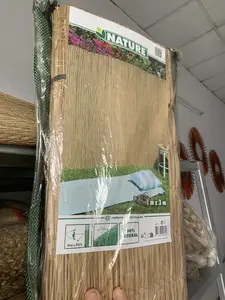 Bức ảnh chụp cận cảnh một cuộn mành che hoặc chiếu được làm bằng vật liệu tự nhiên như cói hoặc sậy, được bọc trong bao bì nhựa trong suốt. Một người đang dùng tay giữ sản phẩm. Bối cảnh dường như là bên trong một cửa hàng hoặc kho hàng, với các sản phẩm dệt tương tự và một chiếc thang ở phía sau. Trên bao bì có in các thông tin chi tiết về sản phẩm bằng tiếng Anh: tên thương hiệu là "NATURE", kích thước "1 m x 3 m", các đặc tính như "100% NATURAL" (100% tự nhiên), "Max. 95%" (mức độ che phủ tối đa), và "±7 mm" (độ dày). Ngoài ra còn có thông tin nhà sản xuất "Ubbink Garden BV, Berenkoog 87, 1822 BN Alkmaar, The Netherlands" và trang web "naturegardening.eu", cho thấy đây là một sản phẩm nhập khẩu.。（日本語訳）。イグサやアシなどの天然素材で作られ、透明なプラスチック包装に包まれた、すだれまたはむしろのロールのクローズアップ写真です。一人の人がその製品を手で持っています。背景は店内または倉庫のようで、後ろには同様の織物製品とはしごが見えます。包装には、製品に関する詳細情報が英語で印刷されています：ブランド名は「NATURE」、サイズは「1 m x 3 m」、特徴として「100% NATURAL」（100%天然）、「Max. 95%」（最大被覆率）、そして「±7 mm」（厚さ）などです。さらに、製造者情報「Ubbink Garden BV, Berenkoog 87, 1822 BN Alkmaar, The Netherlands」とウェブサイト「naturegardening.eu」もあり、これが輸入品であることを示しています。