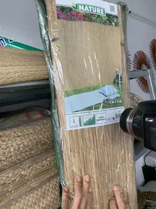 Bức ảnh chụp cận cảnh một người đang dùng tay giữ một tấm mành cuộn, có thể làm từ cói hoặc tre, được bọc trong bao bì ni lông. Bối cảnh dường như là một cửa hàng hoặc kho chứa đồ thủ công mỹ nghệ. Ở phía sau, có thể thấy các sản phẩm đan lát khác, có thể là chiếu cói, được xếp chồng lên nhau trên kệ. Phía bên phải có một chiếc máy ảnh hoặc máy quay phim. Trên bao bì của tấm mành có một nhãn giấy ghi các thông tin: thương hiệu "NATURE", kích thước "1 m x 3 m", và dòng chữ "100% NATURAL". Ngoài ra còn có các thông tin khác như trang web "naturegardening.eu", mã QR, và các biểu tượng mô tả độ che phủ (Max. 95%) và độ dày (± 7 mm) của sản phẩm.。（日本語訳）。ある人が、イグサか竹でできている可能性のある、ビニールで包装された巻すだれを手で持っているクローズアップ写真です。背景は民芸品の店か倉庫のようです。後ろには、棚に積み重ねられた、イグサのござと思われる他の編み物製品が見えます。右側にはカメラかビデオカメラがあります。すだれの包装には、ブランド「NATURE」、サイズ「1m x 3m」、そして「100% NATURAL」という文言の情報が記載された紙のラベルがあります。その他にも、ウェブサイト「naturegardening.eu」、QRコード、そして製品の遮蔽率（最大95%）と厚さ（±7mm）を示すアイコンなどの情報があります。