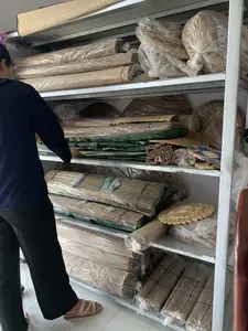 Bối cảnh là một nhà kho hoặc cửa hàng. Một người phụ nữ mặc áo dài tay màu xanh đậm và quần đen đang đứng trước một chiếc kệ sắt nhiều tầng. Người này dường như đang sắp xếp hoặc lựa chọn các sản phẩm. Trên các tầng kệ chứa đầy các sản phẩm thủ công đan lát, chủ yếu là chiếu cói. Các cuộn chiếu được xếp chồng lên nhau, nhiều cuộn được bọc trong bao bì ni lông trong suốt để bảo quản. Ngoài chiếu, còn có các sản phẩm đan lát khác với nhiều hình dạng khác nhau, chẳng hạn như thảm tròn nhỏ và các vật dụng khác cũng được bọc trong túi ni lông. Trên bao bì của một số sản phẩm có dán nhãn nhưng chữ viết không thể đọc được.。（日本語訳）。背景は倉庫か店舗です。濃い青色の長袖と黒いズボンを履いた女性が、多層の鉄製の棚の前に立っています。この人物は、商品を整理しているか、または選んでいるようです。棚の各段は、主にい草のござといった編み物の手工芸品でいっぱいです。巻かれたござは積み重ねられており、多くは保管のために透明なビニール包装で包まれています。ござの他には、小さな円形の敷物やその他の品物など、さまざまな形をした他の編み物製品もあり、それらもビニール袋に包まれています。いくつかの製品の包装にはラベルが貼られていますが、文字は読み取れません。