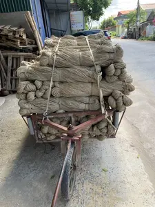 背景は農村地域の村道です。後方には、次のように読める看板があります。「イグサのござ専門生産」。下の行の文字は読みにくいですが、イグサから作られた他の製品がリストされているようです。画像の主な被写体は、道路に停められた手押し車（改良型荷車）で、植物繊維、おそらくイグサの繊維から作られた大きなロープの束が満載されています。これらのイグサ繊維の束は積み重ねられ、ベルトで荷台にしっかりと固定されており、これらはイグサのござのような手工芸品を生産するための原材料です。背景には、民家、木々、そして駐車している車が見えます。画像の左側、屋根の下には、木製パレットの山と竹/笹があります。示されている活動は、伝統的な工芸村のための原材料の輸送です