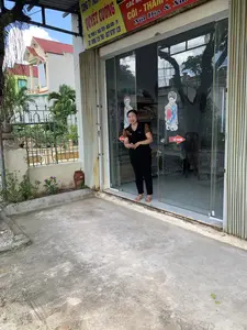 Bức ảnh chụp phía trước một cửa hàng kinh doanh, có thể là một công ty, tại một khu dân cư. Một người phụ nữ trung niên đang đứng ở cửa ra vào bằng kính, đi chân trần và mỉm cười. Bà mặc một chiếc áo màu đen phối nâu và quần dài màu đen.。Phía trên cửa hàng có các tấm biển hiệu. Một tấm biển lớn màu vàng ghi thông tin công ty: 'CÔNG TY TNHH QUYẾT CƯỜNG', 'ĐC: THÔN 3 - NGA TIÊN - NGA SƠN - TH', 'ĐT: 0968 125 768 - 037 9797 128'. Bên cạnh là một tấm biển màu đỏ quảng cáo 'CÁC MẶT HÀNG CÓI - THẢM Nội Địa & Xuất khẩu' (chữ 'MẶT HÀNG' và 'Xuất khẩu' bị che một phần).。Cửa ra vào là loại cửa kính trượt, trên đó có dán hai hình trang trí trẻ em mặc trang phục truyền thống và một miếng dán mũi tên màu đỏ với chữ 'ĐẨY NGANG'. Qua lớp kính, có thể thấy bên trong cửa hàng có các kệ hàng trưng bày sản phẩm, có vẻ là chiếu cói hoặc các sản phẩm thủ công mỹ nghệ.。Không gian phía trước là một khoảng sân xi măng. Bên trái có một hàng rào sắt màu xanh, cây cối và một chậu hoa nhỏ. Phía sau có thể thấy những ngôi nhà khác với mái ngói đỏ.。（日本語訳）。写真は、ある住宅街にある商店、おそらく会社の正面を撮影したものです。一人の中年女性がガラスの出入り口に裸足で立ち、微笑んでいます。彼女は黒と茶色を組み合わせたシャツと黒い長ズボンを着用しています。店の上部には看板があります。大きな黄色い看板には会社の情報が記載されています：「QUYET CUONG有限会社」、「住所：THÔN 3 - NGA TIÊN - NGA SƠN - TH」、「電話番号：0968 125 768 - 037 9797 128」。その隣には、「イグサ製品 - マット 国内＆輸出」と広告する赤い看板があります（「MẶT HÀNG」と「Xuất khẩu」の文字は一部隠れています）。出入り口はガラスの引き戸で、そこには伝統衣装を着た子供の装飾シールが2枚と、「ĐẨY NGANG」（横に押す）という文字が入った赤い矢印のステッカーが1枚貼られています。ガラス越しに、店内には商品を陳列する棚が見え、イグサのござか手工芸品のようです。正面はコンクリートの庭になっています。左側には青い鉄柵、木々、そして小さな植木鉢があります。後方には、赤い瓦屋根の他の家々が見えます。