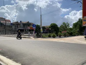 Khung cảnh tại một ngã ba đường nhựa ở một khu dân cư tại Việt Nam. Một người đội mũ bảo hiểm màu vàng đang điều khiển xe máy trên đường. Ở phía sau, có một cây cầu với lan can kim loại. Gần đó có một biển báo màu xanh lam ghi chữ "CẦU HỎI ĐÀO PHÁ NGÃ TIÊN". Dọc theo con đường là dãy nhà phố nhiều tầng, một số đang trong quá trình xây dựng với giàn giáo bao quanh. Có nhiều biển hiệu cửa hàng có thể đọc được, bao gồm "Cửa hàng - ĐỒ GIA DỤNG", "SƠN NHUNG GẠCH DI KIM SƠN", và "THIẾT BỊ VỆ SINH CAO CẤP - SƠN NƯỚC CÁC LOẠI". Một tấm băng rôn quảng cáo treo trên cột điện ghi "GIÁ SỐC 4.990.000đ". Bên phải là một khu vực trồng cây cảnh và hoa trong chậu. Bầu trời có nhiều mây trắng tích tụ.。（日本語訳）。ベトナムのある住宅街のアスファルト道路の三叉路での光景。黄色いヘルメットをかぶった人が道路でバイクを運転している。後方には、金属製の手すりがついた橋がある。その近くには、「CẦU HỎI ĐÀO PHÁ NGÃ TIÊN」と書かれた青い標識がある。道に沿って多層階のタウンハウスが並んでおり、そのうちのいくつかは足場に囲まれて建設中である。「Cửa hàng - ĐỒ GIA DỤNG」、「SƠN NHUNG GẠCH DI KIM SƠN」、「THIẾT BỊ VỆ SINH CAO CẤP - SƠN NƯỚC CÁC LOẠI」など、読み取れる店の看板が多数ある。電柱に掲げられた広告バナーには「GIÁ SỐC 4.990.000đ」と書かれている。右側には、鉢植えの観葉植物や花が植えられたエリアがある。空には白い雲が多く集まっている。