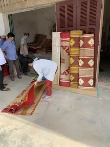 Bối cảnh diễn ra tại một cửa hàng hoặc xưởng bán chiếu cói. Một người phụ nữ đang cúi xuống và trải một chiếc chiếu cói có hoa văn màu đỏ ra nền nhà xi măng. Đứng phía sau cô là hai người đàn ông đang xem xét các sản phẩm. Dựa vào cửa ra vào là những cuộn chiếu cói được cuộn tròn, trưng bày các hoa văn và màu sắc khác nhau như đỏ, vàng và màu tự nhiên của cói. Trên một cuộn chiếu có thể thấy các chữ cái "HIH" được in lặp lại như một phần của hoa văn. Bên trong cửa hàng, có nhiều chồng chiếu khác được xếp gọn gàng. Hoạt động chính là việc mua bán, lựa chọn các sản phẩm chiếu thủ công.。（日本語訳）。舞台は、い草のござを販売する店または工房です。一人の女性がかがみ込み、赤い模様のい草のござをセメントの床に広げています。彼女の後ろには、商品を吟味している二人の男性が立っています。戸口に立てかけられているのは、丸められたい草のござの巻物で、赤、黄、そしてい草の自然な色など、さまざまな模様や色が展示されています。あるござの巻物には、模様の一部として「HIH」という文字が繰り返し印刷されているのが見えます。店内には、他にもたくさんのござがきちんと積み重ねられています。主な活動は、手作りのござ製品の売買や選定です。