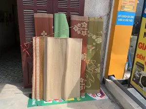 Trong ảnh là cảnh phía trước một ngôi nhà hoặc cửa hàng, nơi những chiếc chiếu dệt được dựng lên để bán. Các chiếc chiếu có nhiều màu sắc và hoa văn khác nhau: một chiếc màu be trơn, một chiếc màu đỏ có hoa văn, một chiếc màu xanh lá cây caro, và một chiếc màu xanh rêu có hoa văn hoa lá. Chúng được dựng dựa vào một cánh cửa chớp bằng gỗ màu nâu đỏ. Bên cạnh là một cây cột màu vàng, trên đó có dán một biển báo màu xanh với dòng chữ "QUÝ KHÁCH VUI LÒNG KHÓA XE CẨN THẬN". Phía trên biển báo là các nhãn dán của các nhà mạng di động như "mobifone", "vietnamobile", "Local", "itec". Một phần của biển quảng cáo máy giặt Panasonic với dòng chữ "XOÁY NƯỚC SIÊU MẠNH" cũng có thể được nhìn thấy ở phía bên phải.。（日本語訳）。写真には、家または店の前の光景が写っており、そこでは織物のござが販売のために立てかけられています。ござには様々な色と模様があります。無地のベージュのもの、模様入りの赤色のもの、緑色のチェック柄のもの、そして花柄のモスグリーンのものです。それらは赤褐色の木製よろい戸に立てかけられています。その隣には黄色い柱があり、そこには「お客様、バイクに慎重に鍵をかけてください」という文言の青い看板が貼られています。看板の上には、「mobifone」、「vietnamobile」、「Local」、「itec」といった携帯通信事業者のステッカーがあります。右側には、「超強力渦水流」という文言が入ったパナソニックの洗濯機の広告看板の一部も見えます。