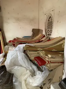 Bên trong một góc phòng, có vẻ là nhà kho hoặc nơi chứa hàng, chứa đầy các sản phẩm thủ công. Các chồng chiếu cói được dệt với nhiều màu sắc và hoa văn khác nhau (đỏ, xanh, màu tự nhiên) được gấp lại và chất thành đống cao. Xen lẫn với chiếu là những chiếc túi ni lông lớn màu trắng và các thùng carton. Bức tường phía sau cũ, có màu trắng ngà và loang lổ. Ở góc tường có một ô cửa sổ nhỏ hình vòm với song sắt trang trí. Trên một thùng carton có thể đọc được chữ "CLAMP MAX 1500" và các biểu tượng.。（日本語訳）。部屋の隅には、倉庫か商品保管場所のようで、手工芸品でいっぱいです。様々な色と模様（赤、青、自然色）で織られたい草のござの山が、折りたたまれて高く積み上げられています。ござに混じって、大きな白いビニール袋と段ボール箱があります。後ろの壁は古く、アイボリーホワイトでまだら模様です。壁の隅にはアーチ型の小さな窓があり、装飾的な鉄格子が付いています。ある段ボール箱の上には「CLAMP MAX 1500」という文字と記号が読み取れます。